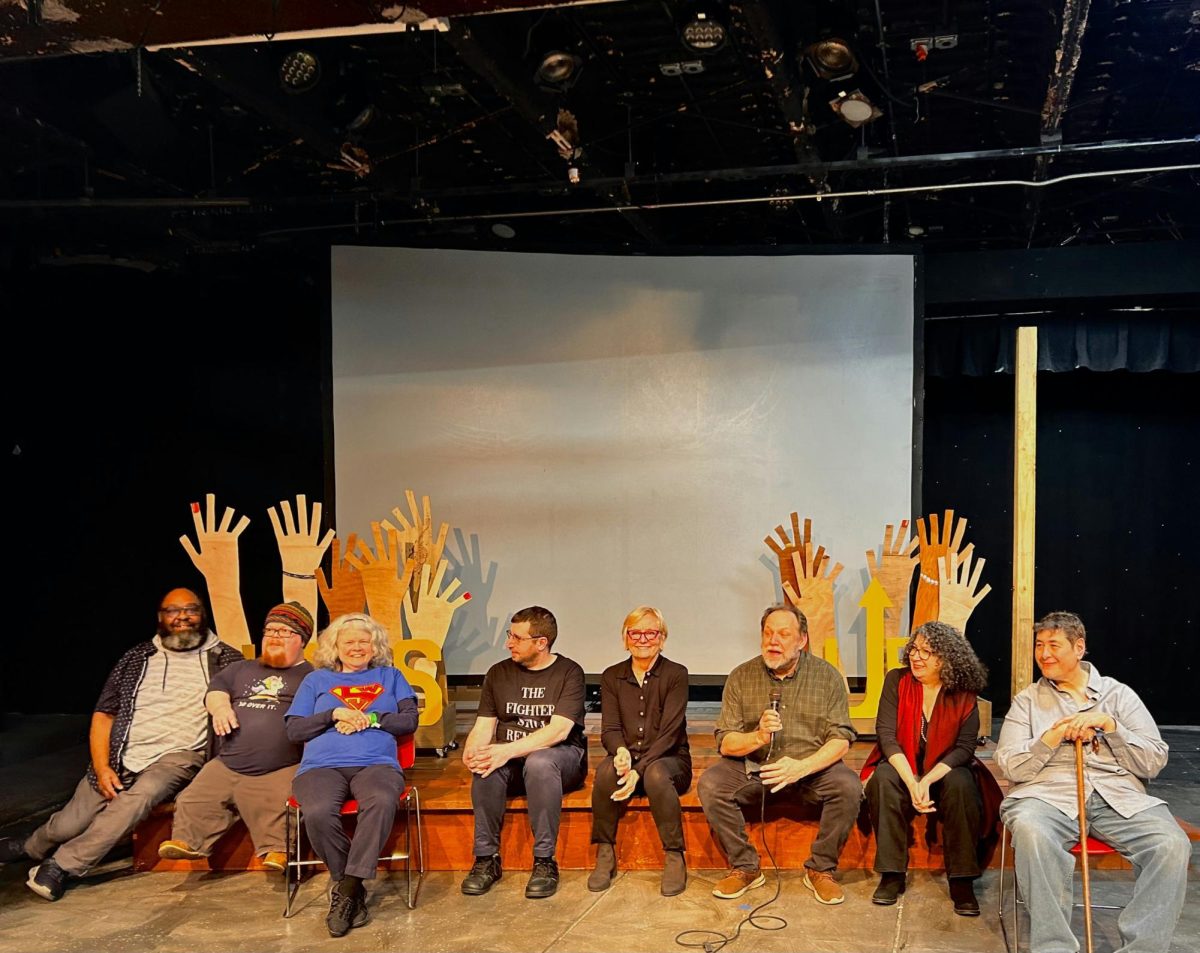 performers are sitting down and smiling during the q&a session with the lights on and the projector behind them is off; Left to Right: Robert Teverbaugh (director),
 Ben Saylor, Mary Hobein, Josh Friedberg, Suzanne Metzel, 
Stephen Donart (director), Joan Afton, Shui Sherard. Stephen Donart is holding a microphone in his right hand.