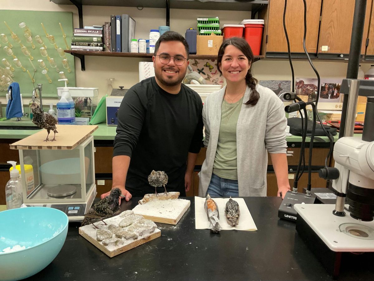  Rafael Sazo, a student (left), stands with Dr. Beth Reinke (right), smiling at the camera. They are in a lab, standing behind a table with 5 taxidermied birds. Three birds are European Starlings posed as if they were alive. On a paper towel, an American Robin lies alongside another European Starling.