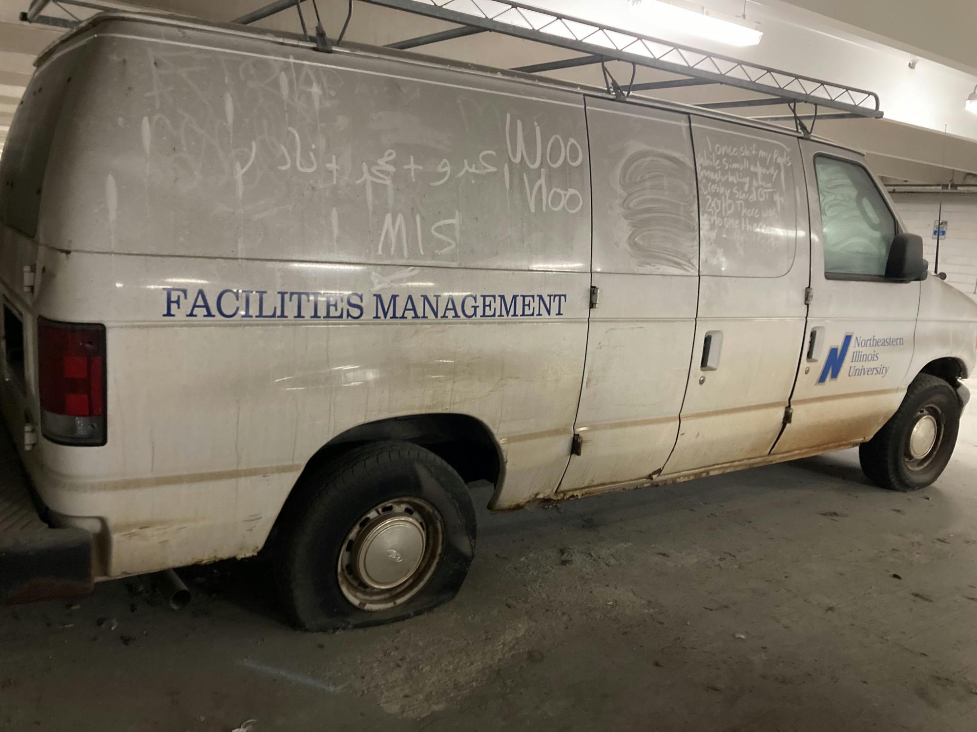 Facilities Management white panel van that was located in the parking garage behind the police station on March 26, 2023. The van in the picture is covered in a visible layer of dust and has a flat tire. Words and pictures have been drawn in the dust on the van. One dust tracing, reportedly made be a BSW, changes the logo to Facilities MisManagement.