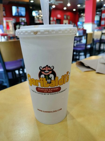 A beverage container with Garibaldi's logo is resting on a table in the restaurant. The logo features a caricature of a smiling Italian man with a long and curly mustache. The blurred background features bright colors and mounted televisions for a luminescent eating atmosphere.