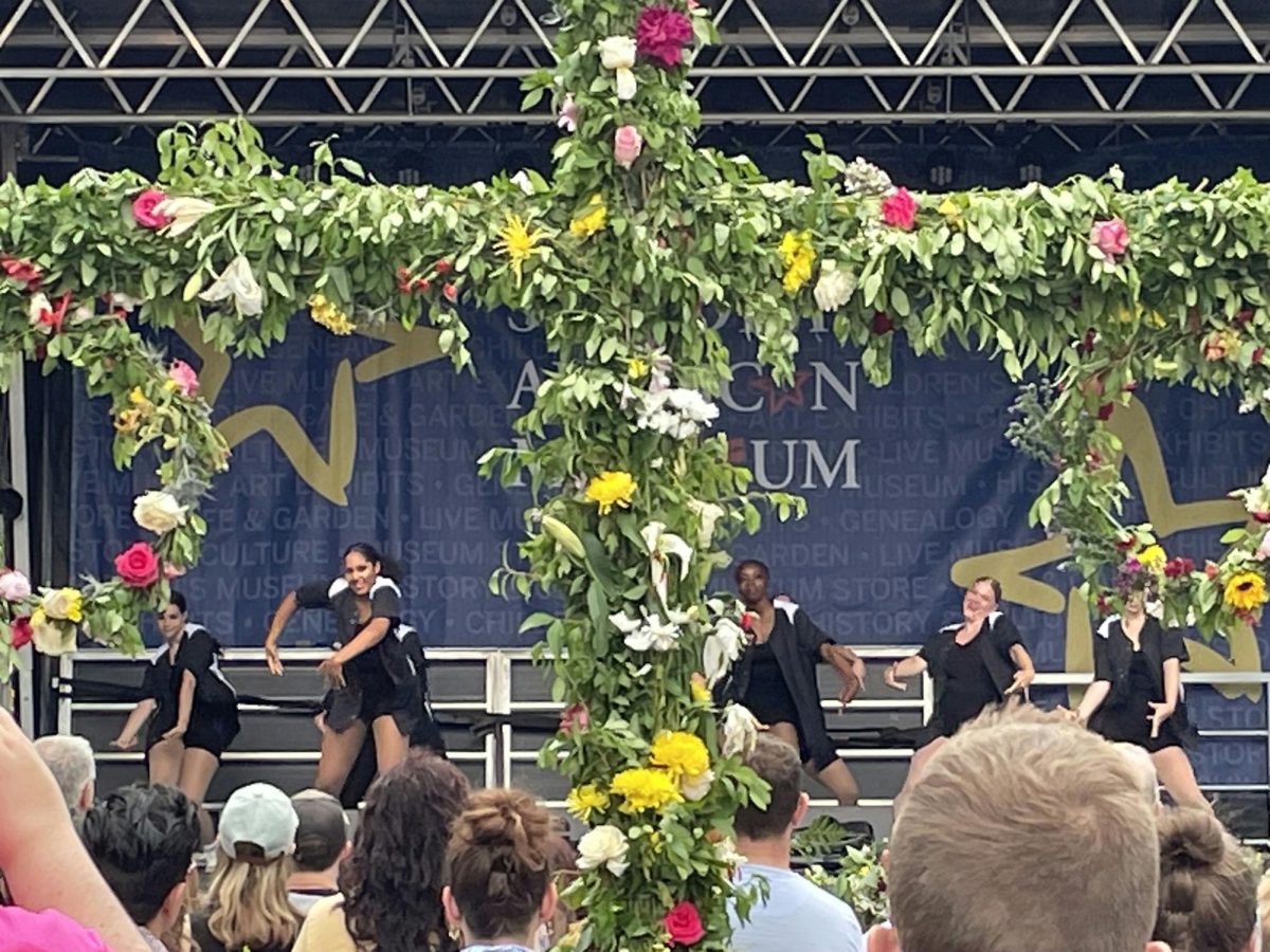 Dancers dressed in black performing on stage in the background with a plant cross is in the foreground, covered with grass and white, yellow and pink flowers.