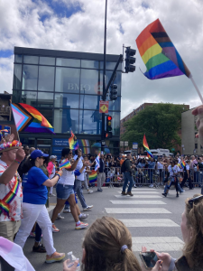 Several people walking down a street, people holding several Pride flags with the inclusion of the trans flag and the black and brown stripes representing People of Color in the shape of the triangle.