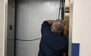 The technician in blue opens the elevator door with a staff member in green smiling as he watches him.