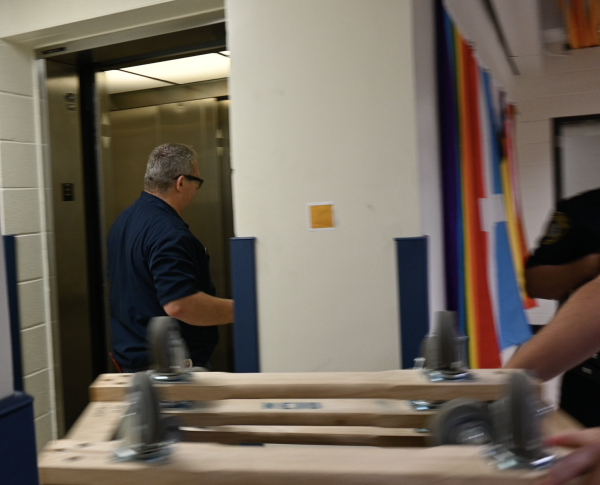 Man in blue uniform standing in front of an open elevator with a dolly rolling past in the foreground