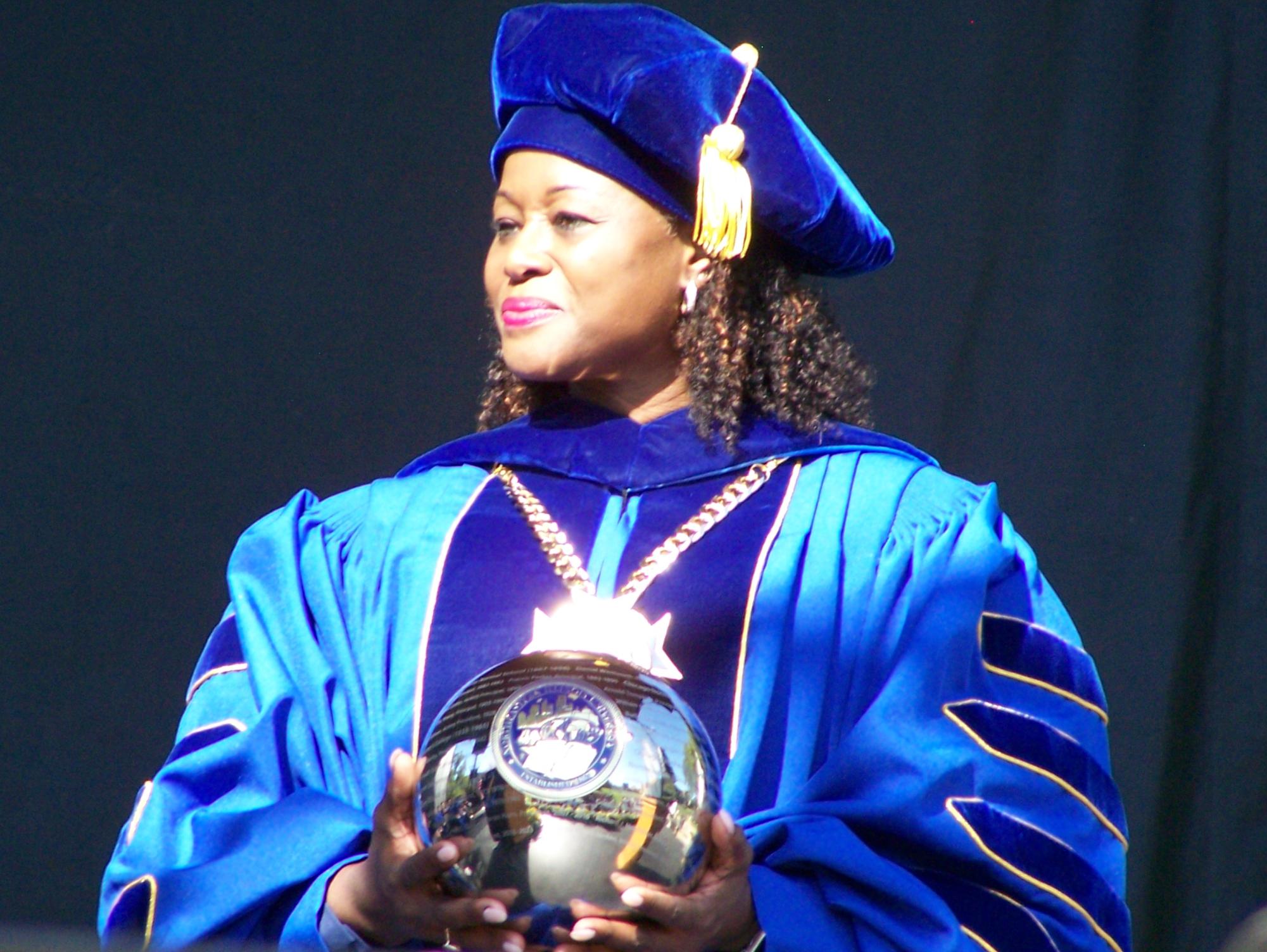 President Dr. Bell Jordan In blue regalia wearing the presidential medallion. She is looking to the side holding the University Orb