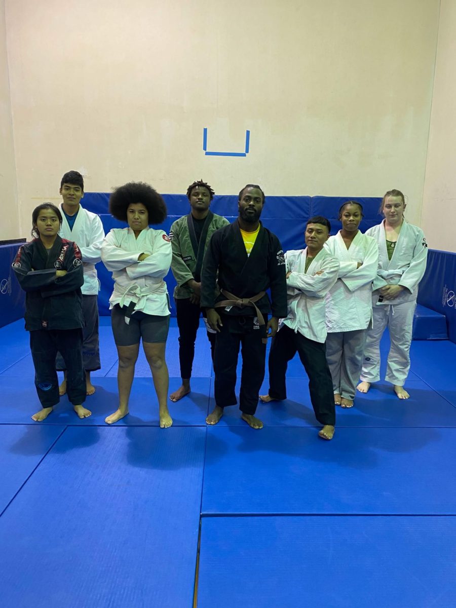 Eight members of NEIU’s 2025 Jiu Jitsu Class are posing for a photographer in their traditional garments called a gis, and each of them is wearing a different colored belt that symbolizes a different level of expertise. 