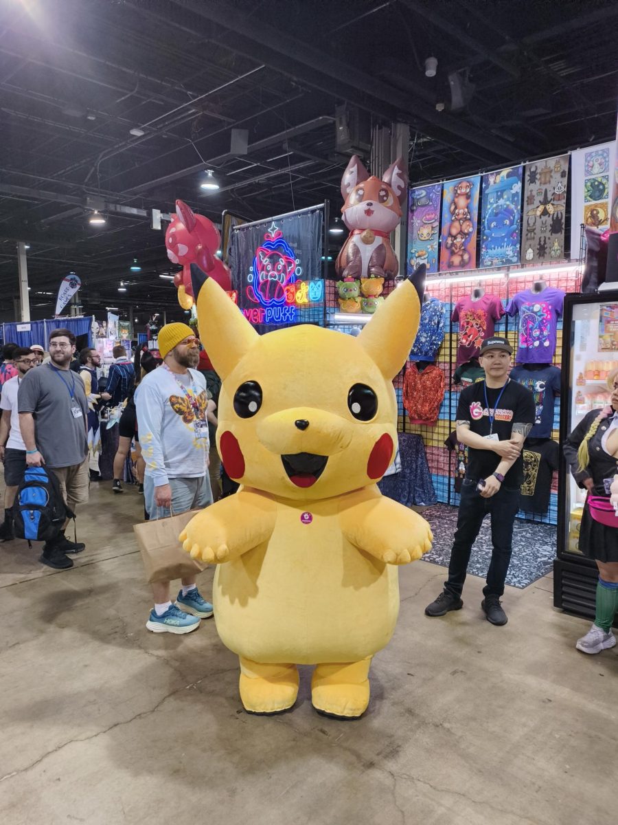 A person in a Pokemon Pikachu costume is posing for the photo with a booth and ACen visitors in the background.